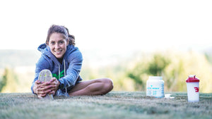 Young woman stretching leg on grass with a tub of Goat Whey and a shaker bottle